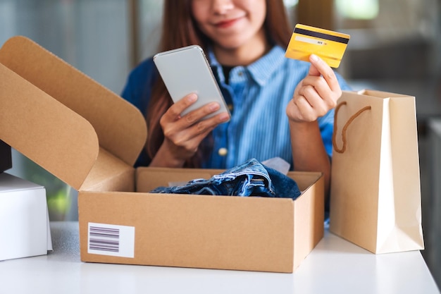 Foto close-up beeld van een mooie jonge vrouw die mobiele telefoon en creditcard gebruikt voor online winkelen met boodschappentas en postpakketdoos met kleding op tafel
