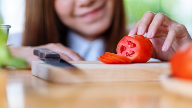 Close-up beeld van een mooie vrouw die tomaat kijkt en snijdt met een mes op een houten bord