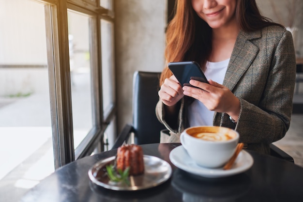 Close-up beeld van een vrouw die mobiele telefoon vasthoudt en gebruikt met koffiekopje en snack op tafel in café