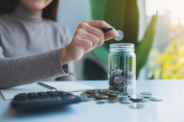 Close-up beeld van een vrouw die munten in een glazen pot stopt met rekenmachine op tafel om geld en financieel concept te besparen