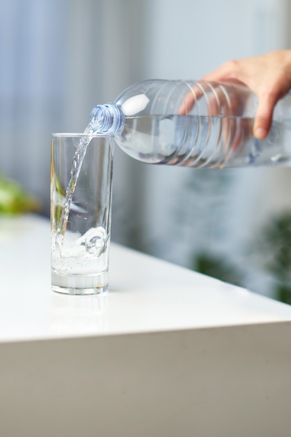 Close-up beeld van een vrouwelijke hand die drinkwaterfles vasthoudt en water in glas op tafel giet.
