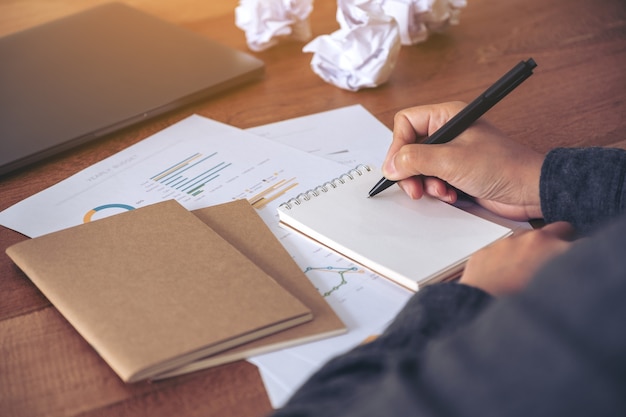 Close-up beeld van een zakenvrouw werken en opschrijven op een witte lege notebook met verpest papieren en laptop op tafel in kantoor