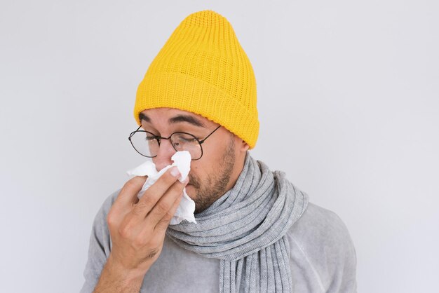 Close-up beeld van zieke knappe man met grijze trui gele hoed en bril neus snuiten en niezen in weefsel Man heeft griepvirus of allergie luchtwegen Gezonde geneeskunde en mensen concept