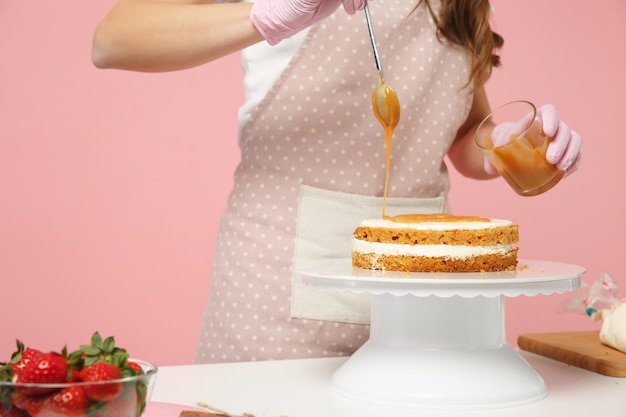 Foto close-up bijgesneden chef kok banketbakker of bakker in wit t-shirt koken aan tafel geïsoleerd op roze pastel achtergrond in studio. crème applicatie cake maken proces. bespotten kopie ruimte voedsel concept.