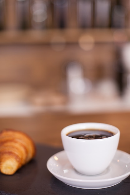 Close up of black coffee next to freshly backed croissant on the bar counter.