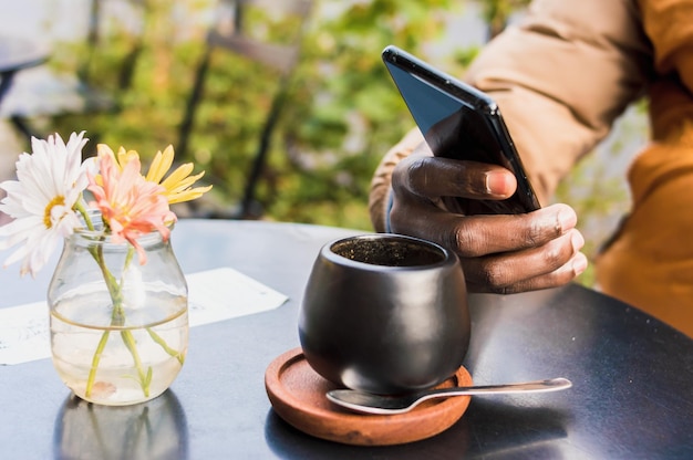 Chiuda sulla mano maschio nera che tiene un telefono su un tavolo con una tazza di caffè che si siede all'aperto tecnologia