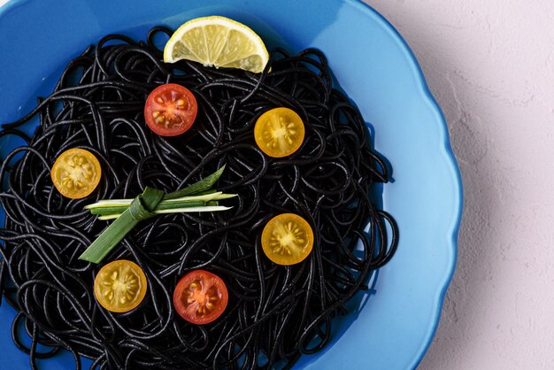 Close-up of black pasta with cuttlefish ink with yellow and red tomatoes in blue plate on pink background, vegetarian food, time to eat concept