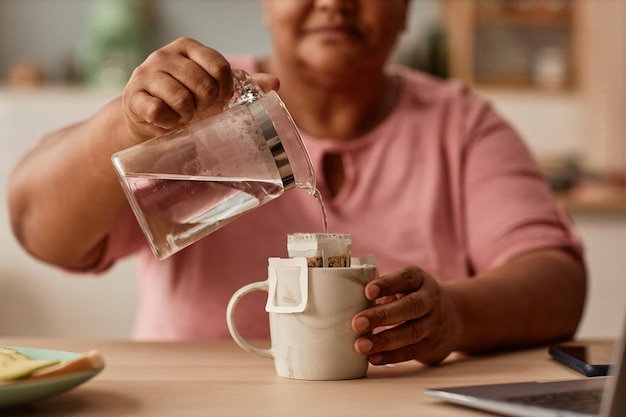 Close up of black senior woman making tea in cozy home kitchen holding glass kettle and pouring wate