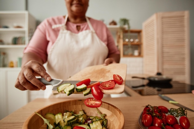 Photo close up of black senior woman putting vegetables in bowl while making salad in kitchen copy space