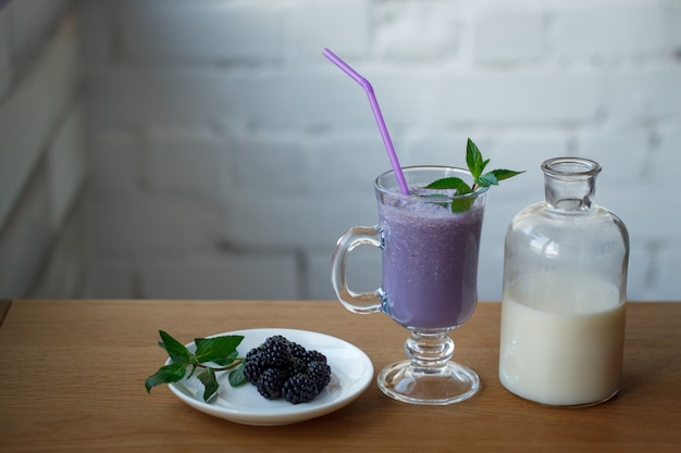 Frullato di more in una tazza di vetro, una bottiglia di latte e un piattino con bacche fresche e un rametto di menta