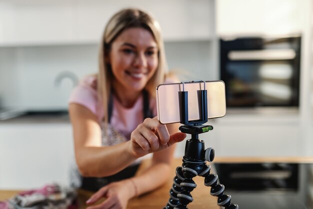 Close-up of blond housewife using smart phone on tripod in a kitchen for searching recipe.