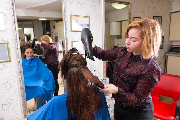 살롱에서 브러시와 핸드 블로우 건조기를 사용하여 젊은 여성 갈색 머리 클라이언트의 머리를 건조하는 금발 스타일리스트의 클로즈업