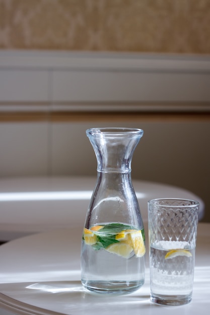 Close-up of a bottle with a cap and a glass of water with mint and lemonon a white table and sun glare