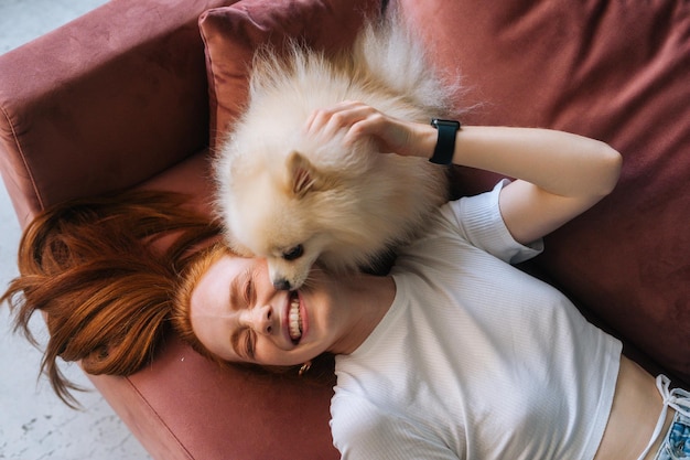 Close-up bovenaanzicht van mooie witte kleine Spitz-huisdierhond, heerlijk likkend gezicht van lachende jonge vrouw die op een comfortabele bank ligt. Aantrekkelijke roodharige vrouw die vrije tijd doorbrengt met pluizig hondje thuis.