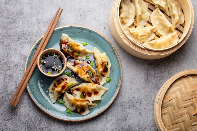 Close-up, bovenaanzicht van traditionele Aziatische/Chinese dumplings in blauw bord met sojasaus, eetstokjes en een bamboestoomboot op grijze rustieke stenen achtergrond. Authentieke Chinese keuken