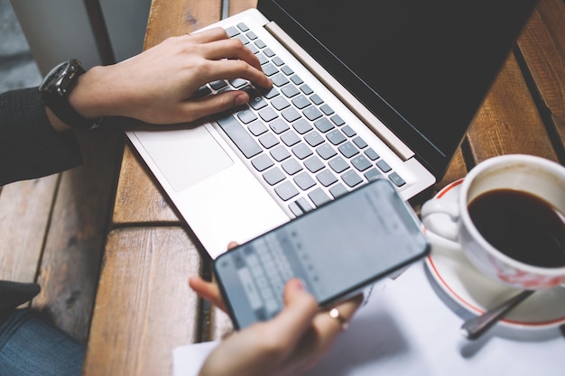 Close-up bovenaanzicht van vrouwelijke handen met smartphone met groot scherm vooraan laptop toetsenbord werkend