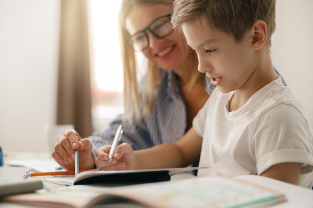 Close up of boy sitting at table and writing in copybook on online lesson while his mom helping him