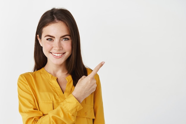Photo close-up of brunette female model showing promo offer, pointing finger right and smiling at front, white wall.