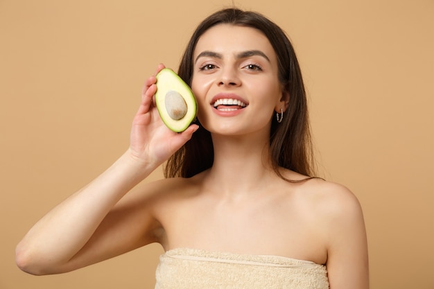 Close-up brunette half naakte vrouw met perfecte huid naakt make-up houdt avocado geïsoleerd op beige pastel muur
