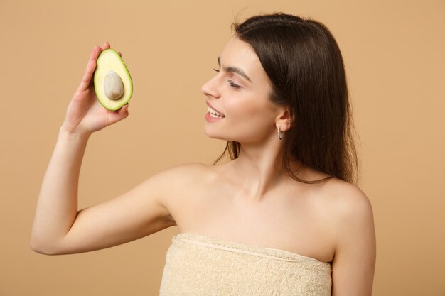 Close-up brunette half naakte vrouw met perfecte huid naakt make-up houdt avocado geïsoleerd op beige pastel muur