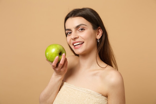 Close up brunette half naked woman with perfect skin, nude make up holds apple isolated on beige pastel wall