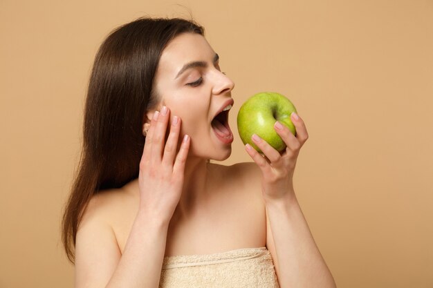 Close up brunette half naked woman with perfect skin, nude make up holds apple isolated on beige pastel wall