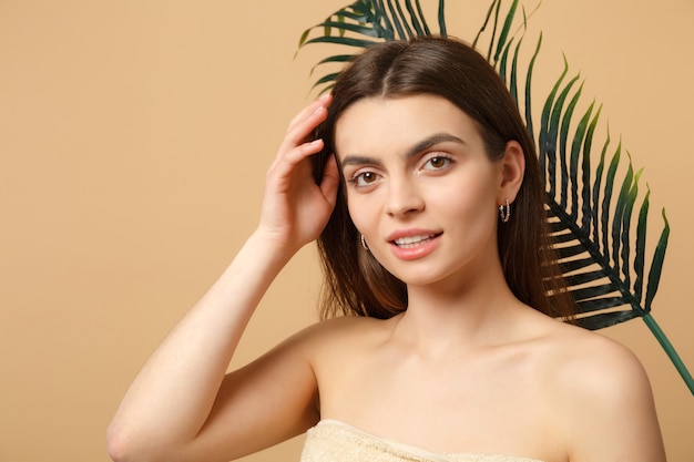 Close up brunette half naked woman with perfect skin, nude make up and palm leaf isolated on beige pastel wall