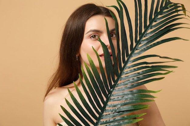 Close up brunette half naked woman with perfect skin, nude make up and palm leaf isolated on beige pastel wall