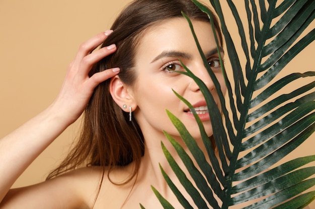 Close up brunette half naked woman with perfect skin, nude make up and palm leaf isolated on beige pastel wall