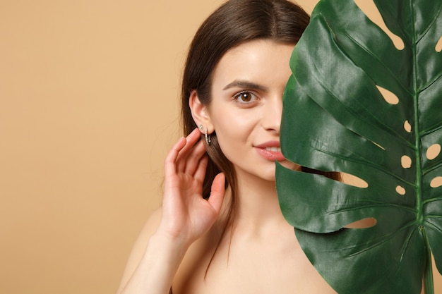 Close up brunette half naked woman with perfect skin, nude make up and palm leaf isolated on beige pastel wall