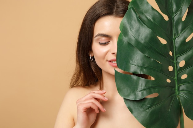 Close up brunette half naked woman with perfect skin, nude make up and palm leaf isolated on beige pastel wall