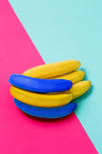 Close up of a bunch of bananas on a colored pink and blue background. Two bright blue fruits are among the yellow