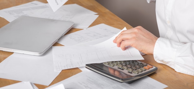 Close up of businessman or accountant hand holding pen working on calculator accountancy document and laptop computer at office, business concept