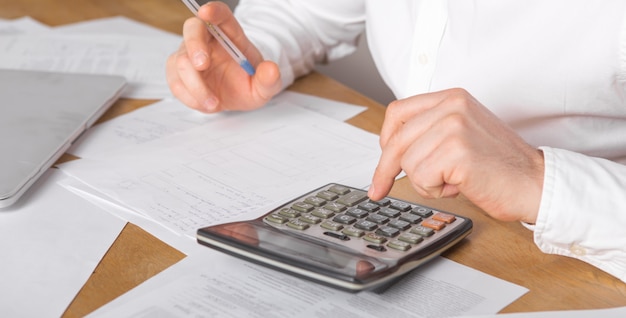 Close up of businessman or accountant hand holding pen working on calculator accountancy document and laptop computer at office, business concept