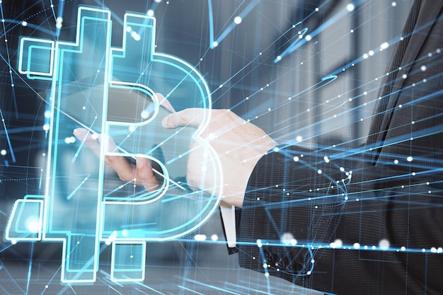 Close up of businessman hand pointing at tablet with abstract glowing holographic bitcoin icon on blurry office interior background Cryptocurrency online banking and money concept Double exposure