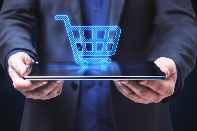 Close up of businessman hands holding tablet with abstract shopping cart on blue background Ecommerce and online shopping concept