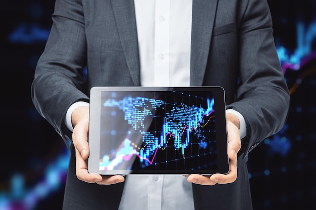 Close up of businessman hands holding tablet with glowing candlestick forex chart map hologram on dark background Trade and market concept