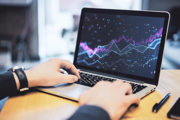 Close up of businessman hands using laptop keyboard with abstract glowing candlestick forex chart with index and grid on modern workplace Invest trade finance ans stock market concept