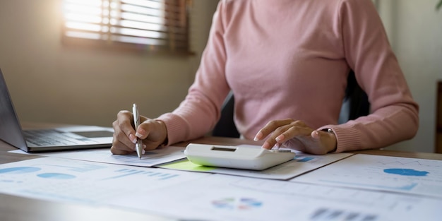 Close up of businesswoman or accountant hand holding pen working on calculator to calculate business data accountancy document and laptop computer at office business concept