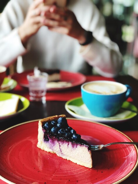 Photo close-up of cake slice in plate with person using mobile phone in background on table