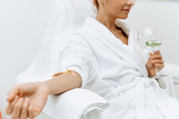 Photo close up of calm female patient sitting with tube and needle during iv infusion. girl holding glass with lemon water and relaxing