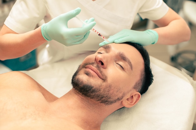 Close up of the calm young man lying with his eyes closed and hands of the cosmetologist touching his forehead and making hyaluronic acid injection