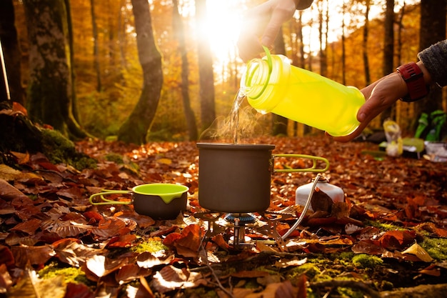 Close-up campingpan verwarmd op draagbaar gasfornuis Man's hand gieten water in de pan van gele fles Heerlijk herfstbos op een wazige achtergrond Elk seizoen is geschikt om te wandelen