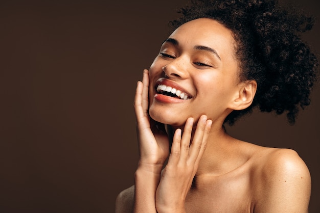 Close up of carefree young woman laughing. Portrait of smiling woman with naked shoulders and closed eyes smiling while posing. Beautiful girl isolated on brown background