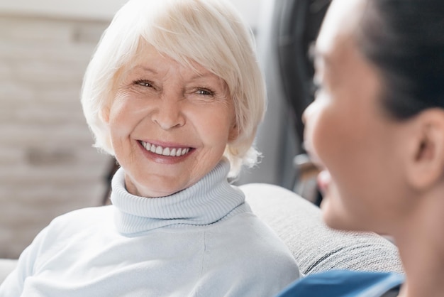 Photo close up of caregiver smiling and talking to aged woman while taking care of her at home