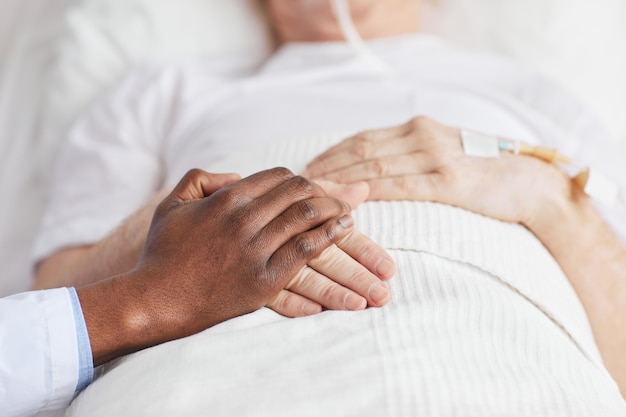 Primo piano di un medico afroamericano premuroso che si tiene per mano con un paziente anziano sdraiato nel letto d'ospedale, copia spazio