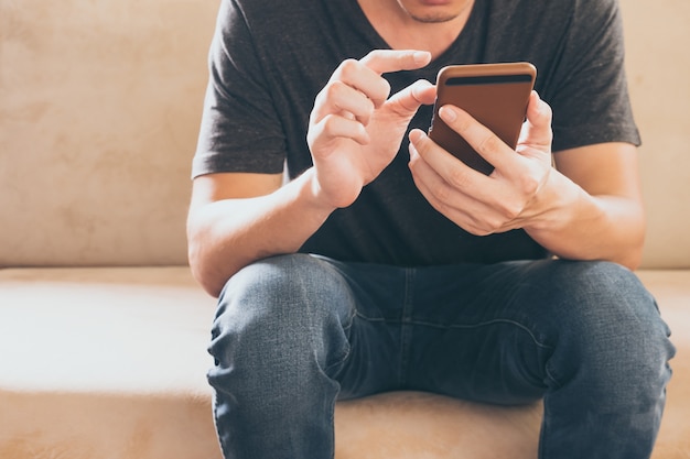 Close up of casual young man sitting on sofa and usig a smart phone at home.