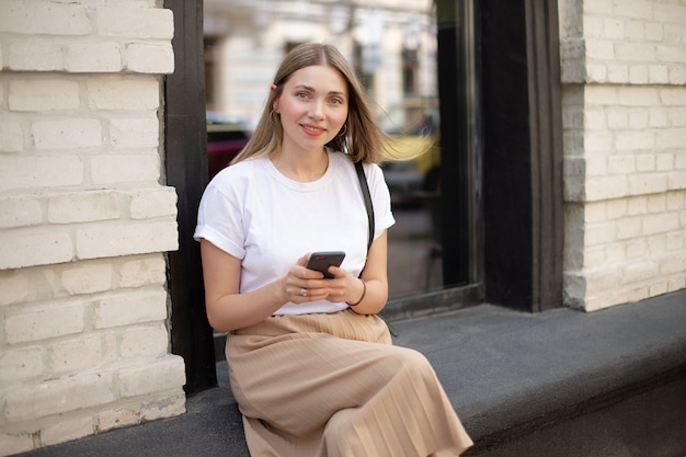 Primo piano della ragazza bionda caucasica in una t-shirt bianca utilizzando lo smartphone mentre è seduto per strada sullo sfondo di una grande finestra vicino a un edificio in mattoni