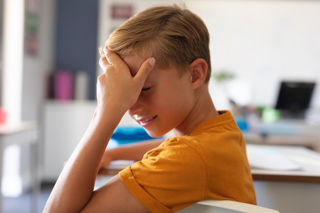 Close-up of caucasian elementary schoolboy with head in hand sitting at desk in classroom