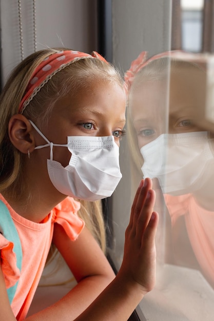 Close-up of caucasian elementary schoolgirl wearing mask while looking through window